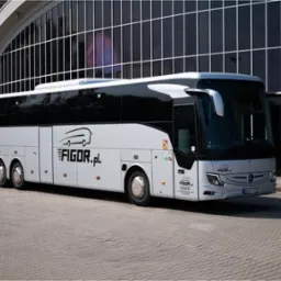 Nowoczesny, srebrny autokar z czarnymi oknami i logo firmy Figor.pl, zaparkowany przed budynkiem z ciemnymi, szklanymi panelami.