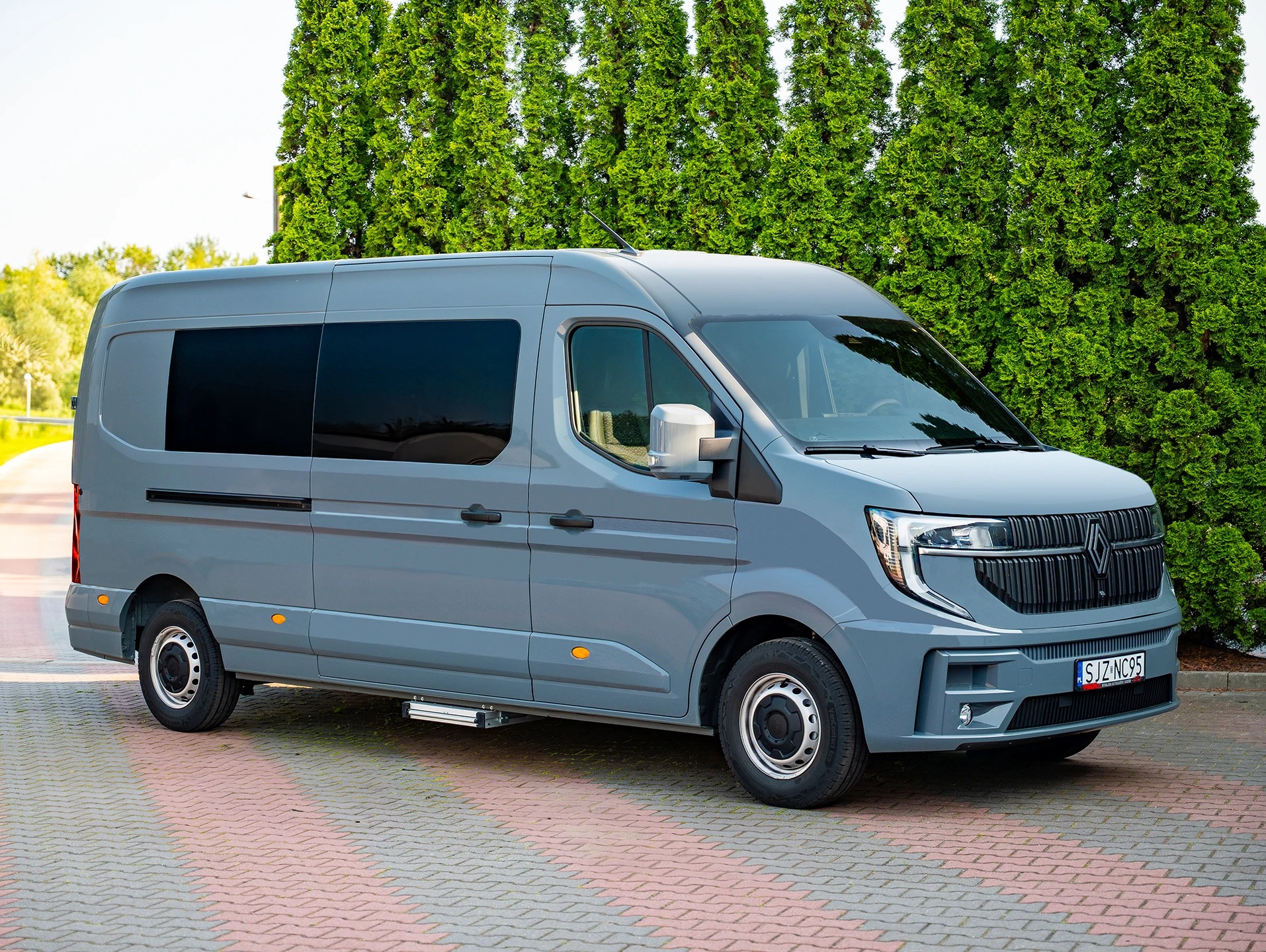 Szary bus na tle zielonych drzew. Nowoczesny design, aluminiowy stopień. Charakterystyczny grill i reflektory. Pojazd zaparkowany na kostce brukowej.