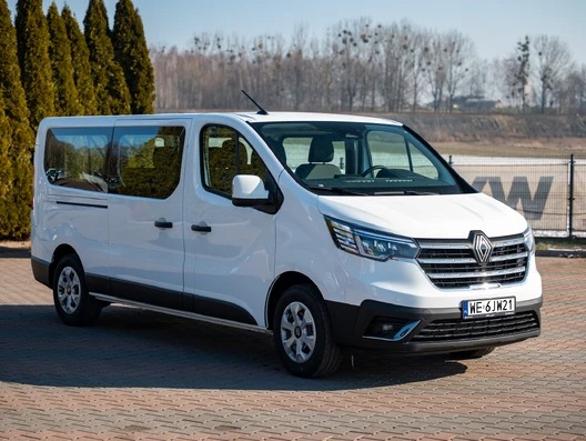 Biały, ośmioosobowy bus Renault Trafic z podłużnymi oknami i rejestracją WE 6JW21, zaparkowany na brukowanym placu w słoneczny dzień.
