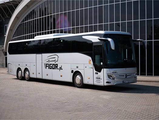Nowoczesny, srebrny autokar z czarnymi oknami i logo firmy Figor.pl, zaparkowany przed budynkiem z ciemnymi, szklanymi panelami.