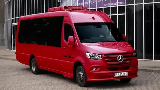 Czerwony, lśniący bus Mercedes-Benz Sprinter z czarnymi szybami na tle nowoczesnego budynku z metalowymi elementami. Widoczny z przodu, lekko z boku.