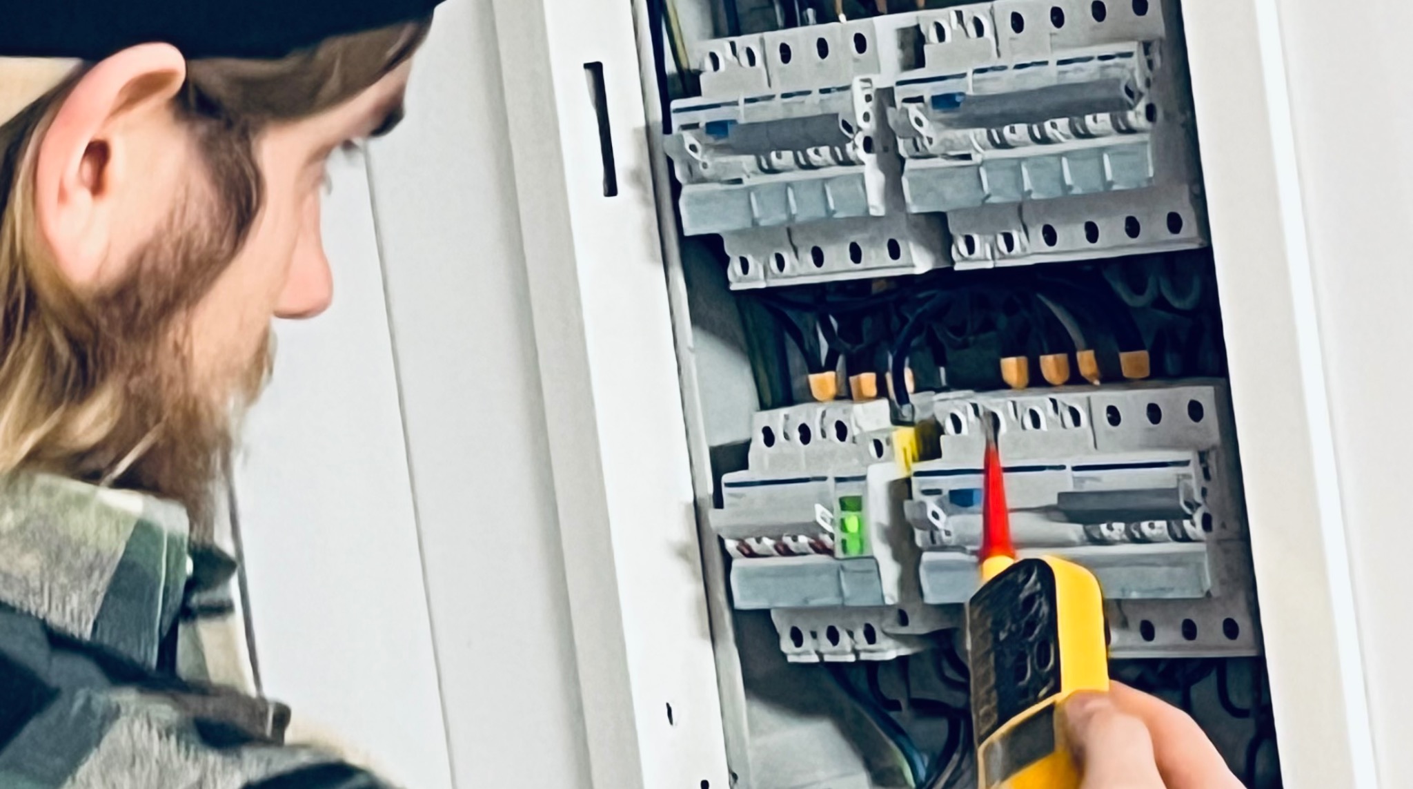 Elektryk z brodą sprawdza napięcie w skrzynce bezpiecznikowej za pomocą próbnika. Widoczne bezpieczniki, przewody i elementy instalacji elektrycznej.