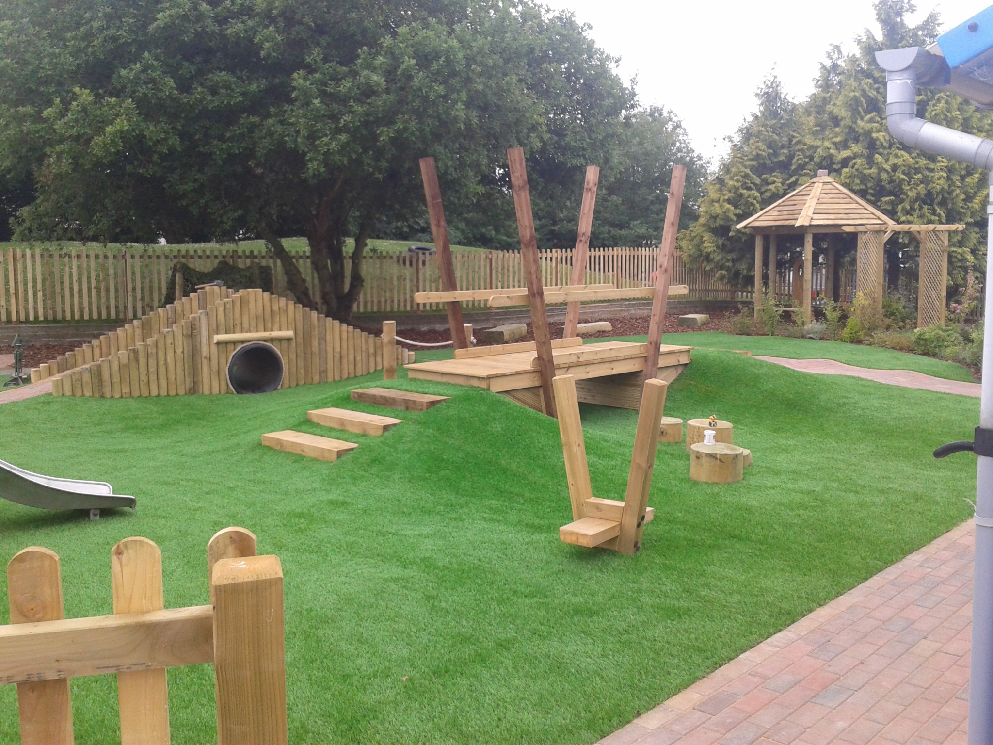 Kompleksowy plac zabaw z drewna: tunel, mostek, zjeżdżalnia i altana. Elementy osadzone na sztucznej trawie, otoczone zielenią i ogrodzeniem.