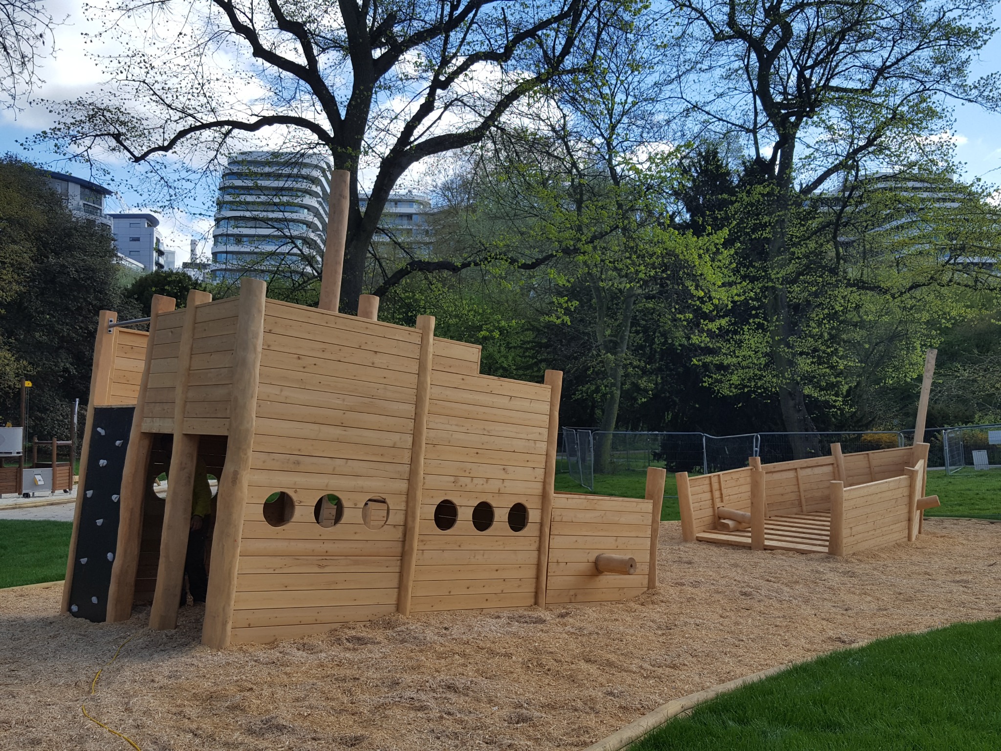 Drewniany plac zabaw w kształcie statku z elementami wspinaczkowymi na tle zieleni miejskiej i nowoczesnego budynku. Jasne drewno, okrągłe okna, piaskowe podłoże.