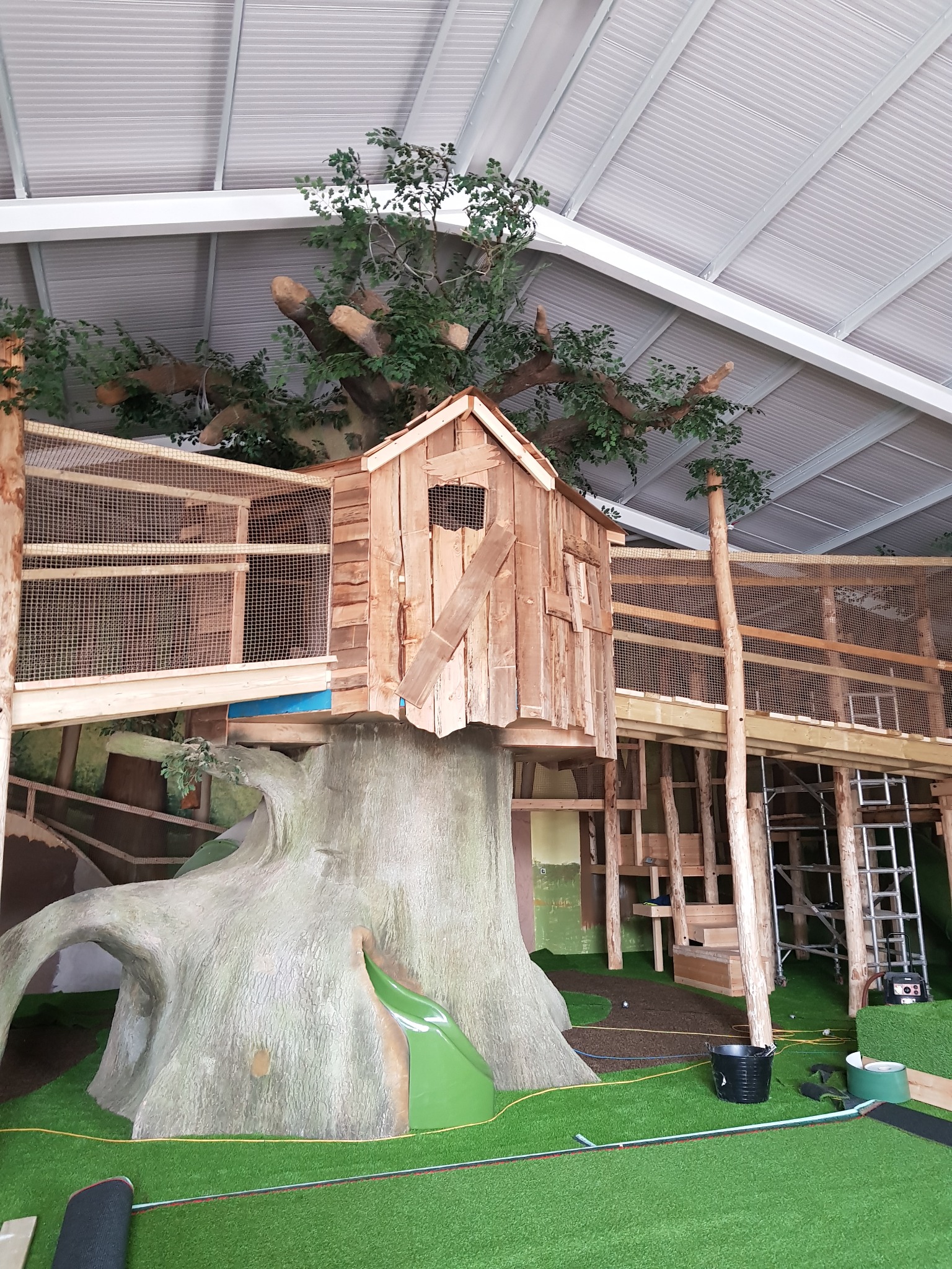 Rozbudowany plac zabaw w Bolesławcu: drewniany domek na sztucznym drzewie z mostkami i zjeżdżalnią, całość pod zadaszeniem. Widoczne elementy konstrukcyjne i narzędzia.