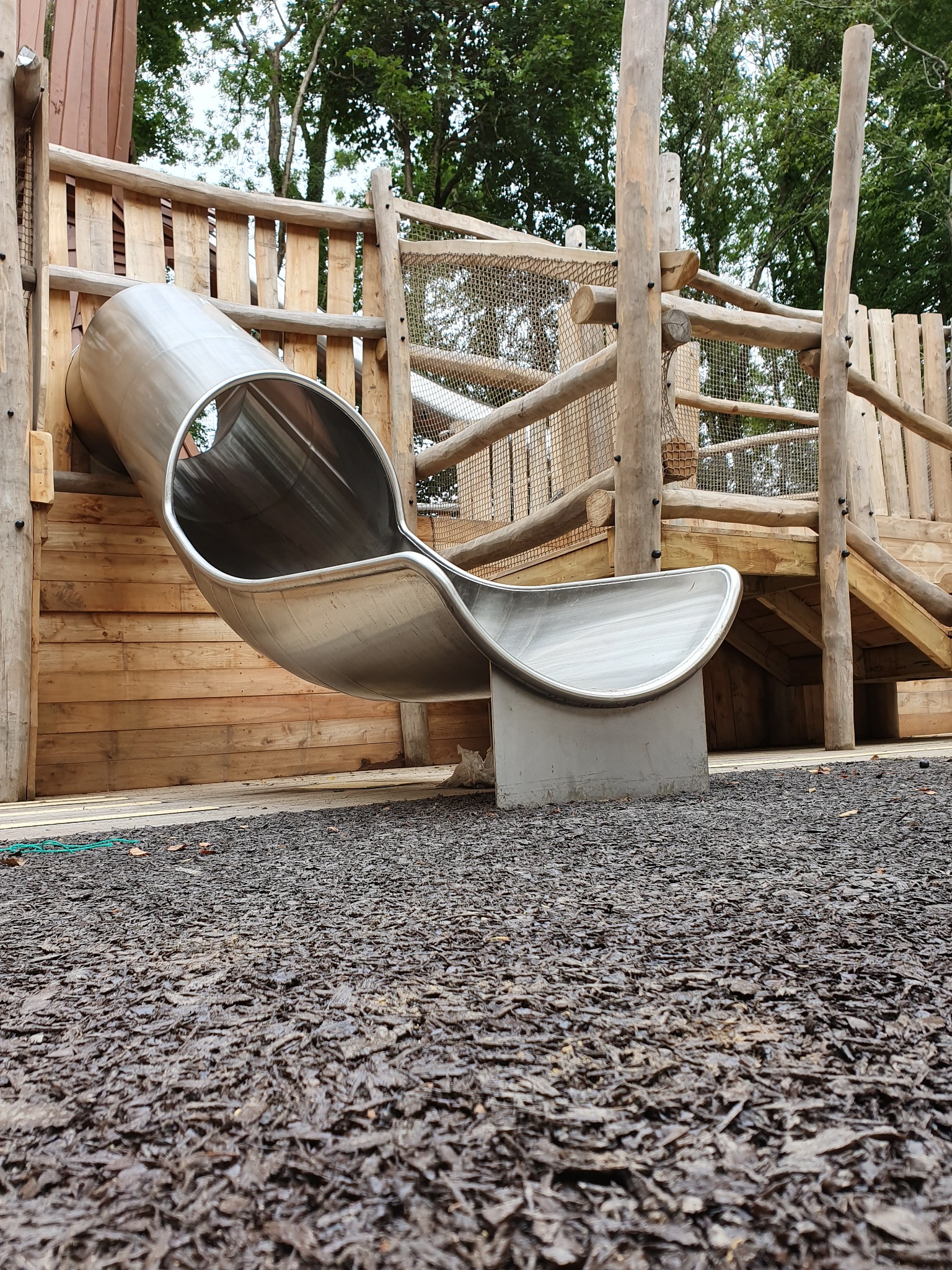 Metalowa zjeżdżalnia na drewnianym placu zabaw z siatką zabezpieczającą, widziana z perspektywy gruntu pokrytego korą.