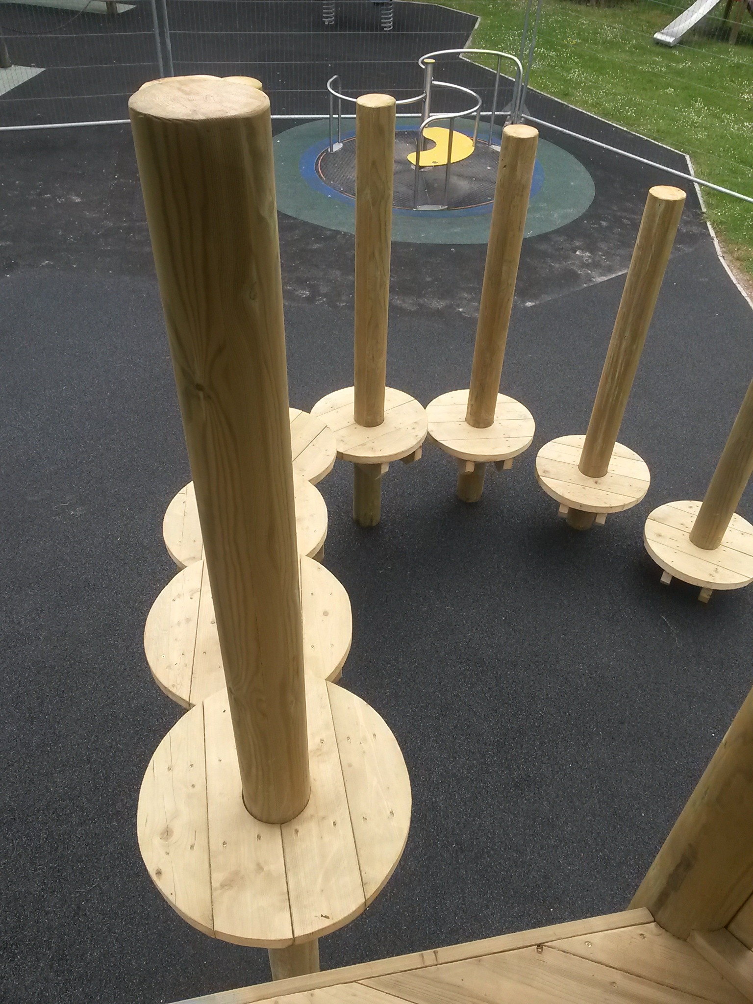 Drewniany tor przeszkód na placu zabaw z okrągłymi platformami i pionowymi słupami, widok z góry, bezpieczna nawierzchnia, elementy metalowe w tle.