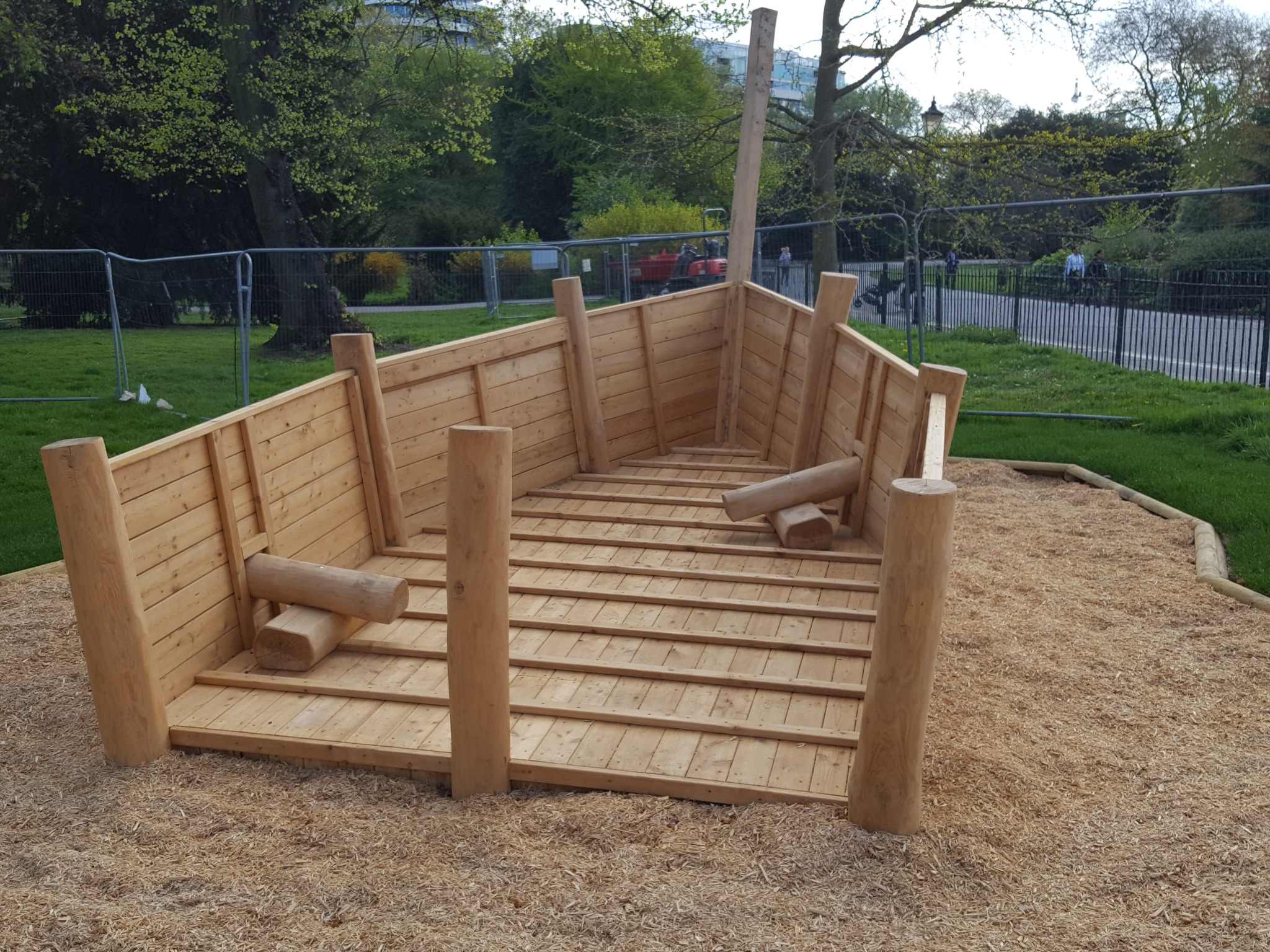 Drewniana konstrukcja w kształcie łodzi na placu zabaw, z elementami do wspinaczki i zabawą w piasku. W tle zieleń parku i ogrodzenie.