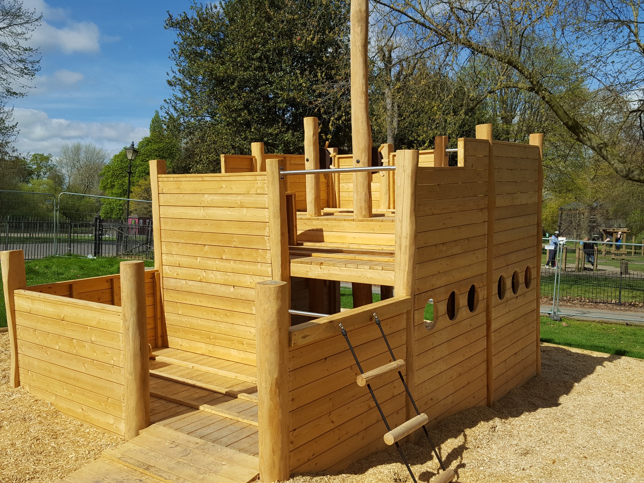 Drewniany plac zabaw w kształcie statku z drabinką sznurową i otworami okiennymi, na tle zieleni parku i błękitnego nieba.