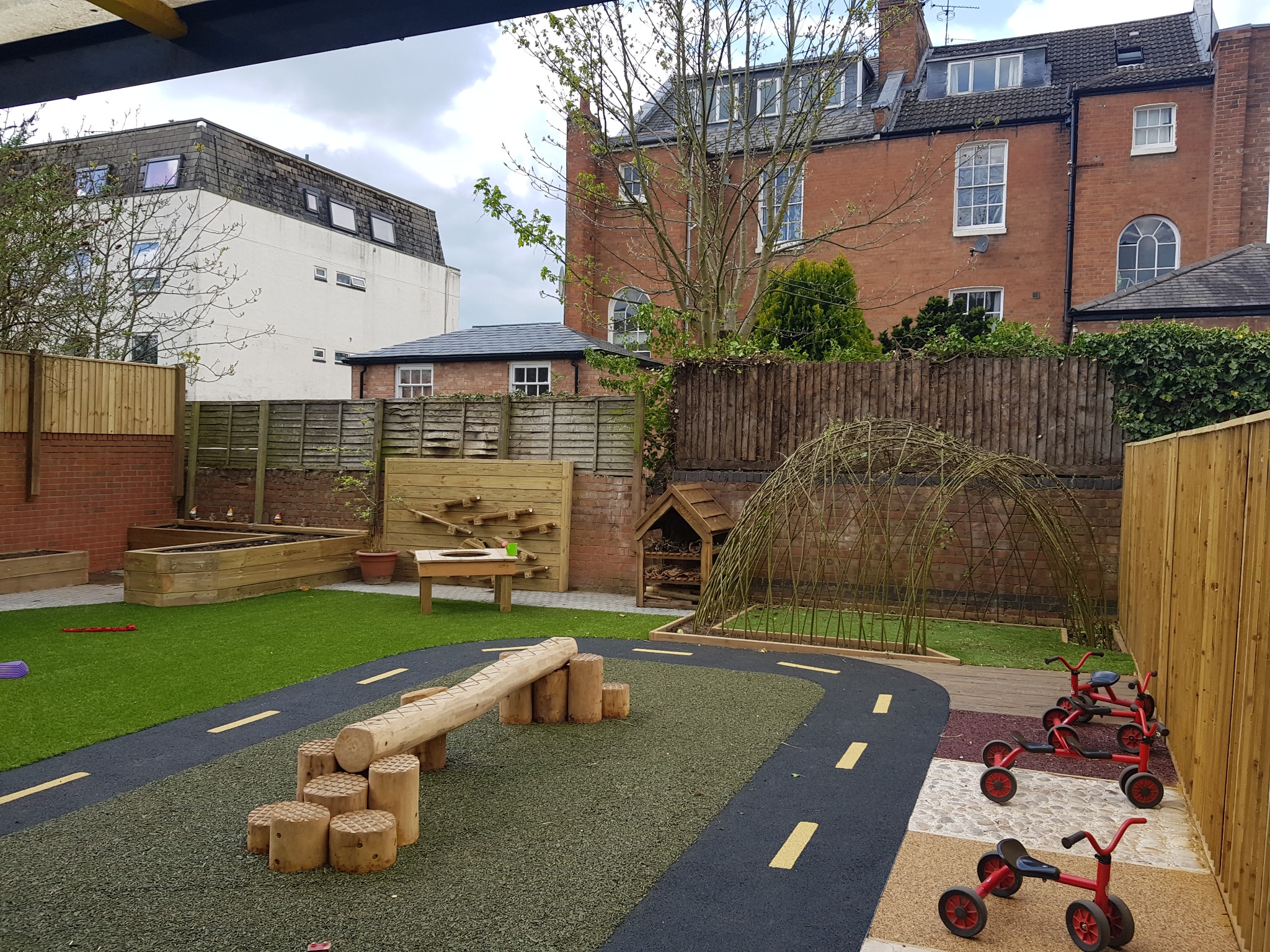 Ukończony plac zabaw z drewnianymi elementami: ścieżka z przeszkodami, domek, ścianka wspinaczkowa i trójkołowe rowerki. Trawa, ogrodzenie i budynki w tle.