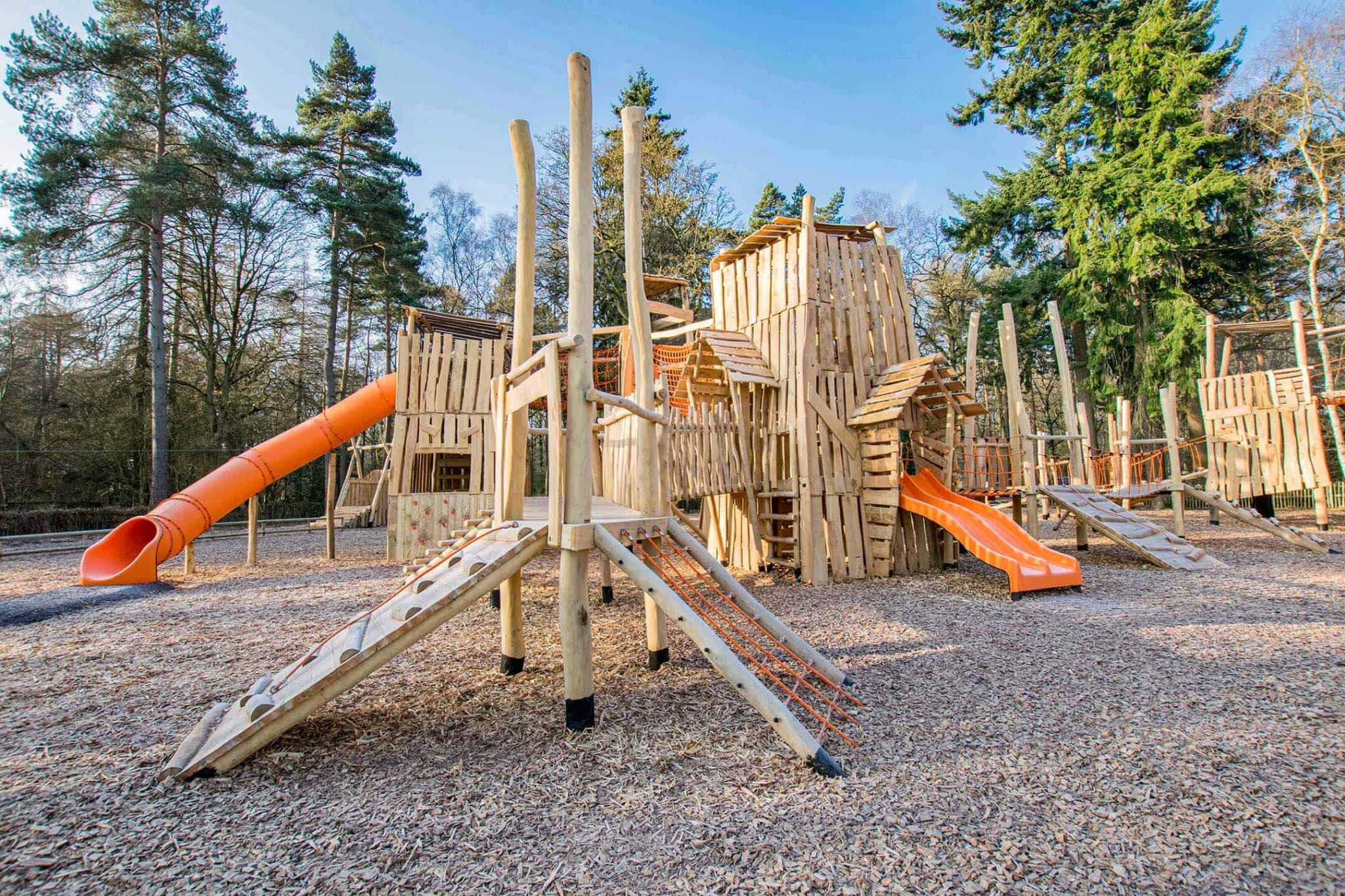 Rozległy, drewniany plac zabaw z wieżami, zjeżdżalniami i mostkami linowymi, otoczony drzewami i pokryty korą drzewną. Jasne, naturalne światło.