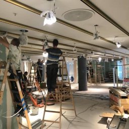 Sonicelektro - Montaż oświetlenia sufitowego w dużej przestrzeni. Elektrycy na drabinach instalują lampy w osłonach, widoczne przewody i narzędzia.