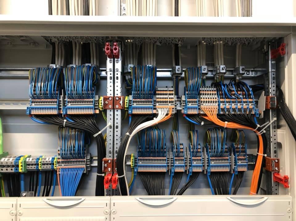 Rozdzielnica elektryczna z precyzyjnie ułożonymi przewodami w różnych kolorach, uporządkowanymi w wiązki i podłączonymi do listew zaciskowych. Widoczne elementy montażowe i kable w peszlu.