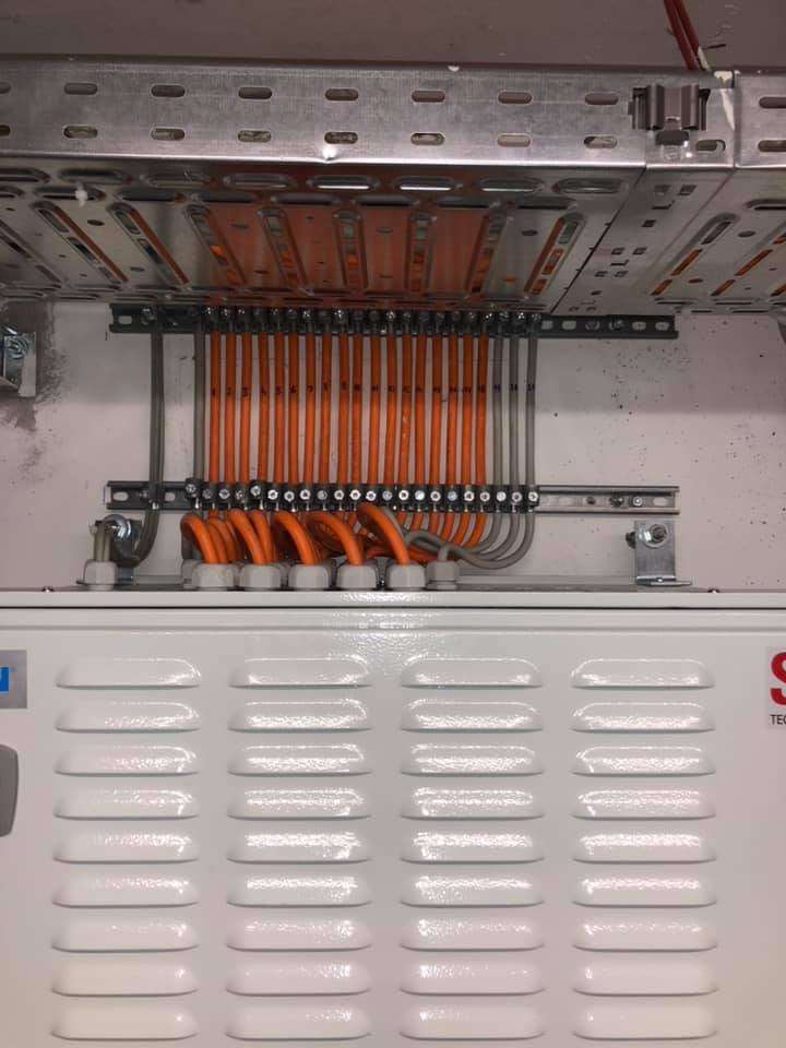 Szczegółowe ujęcie rozdzielnicy elektrycznej z pomarańczowymi kablami w metalowej korycie, widoczne elementy mocujące i wentylacja obudowy.