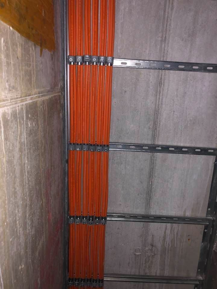 Równolegle ułożone pomarańczowe przewody elektryczne, przymocowane do metalowych szyn na surowej ścianie. Widoczne mocowania i fragment ściany z teksturą betonu.