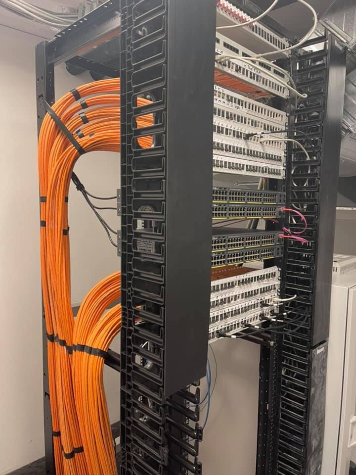 Zorganizowany panel krosowniczy z pomarańczowymi kablami sieciowymi w szafie rack, widoczne przełączniki i patch panele. Estetyczne ułożenie przewodów.