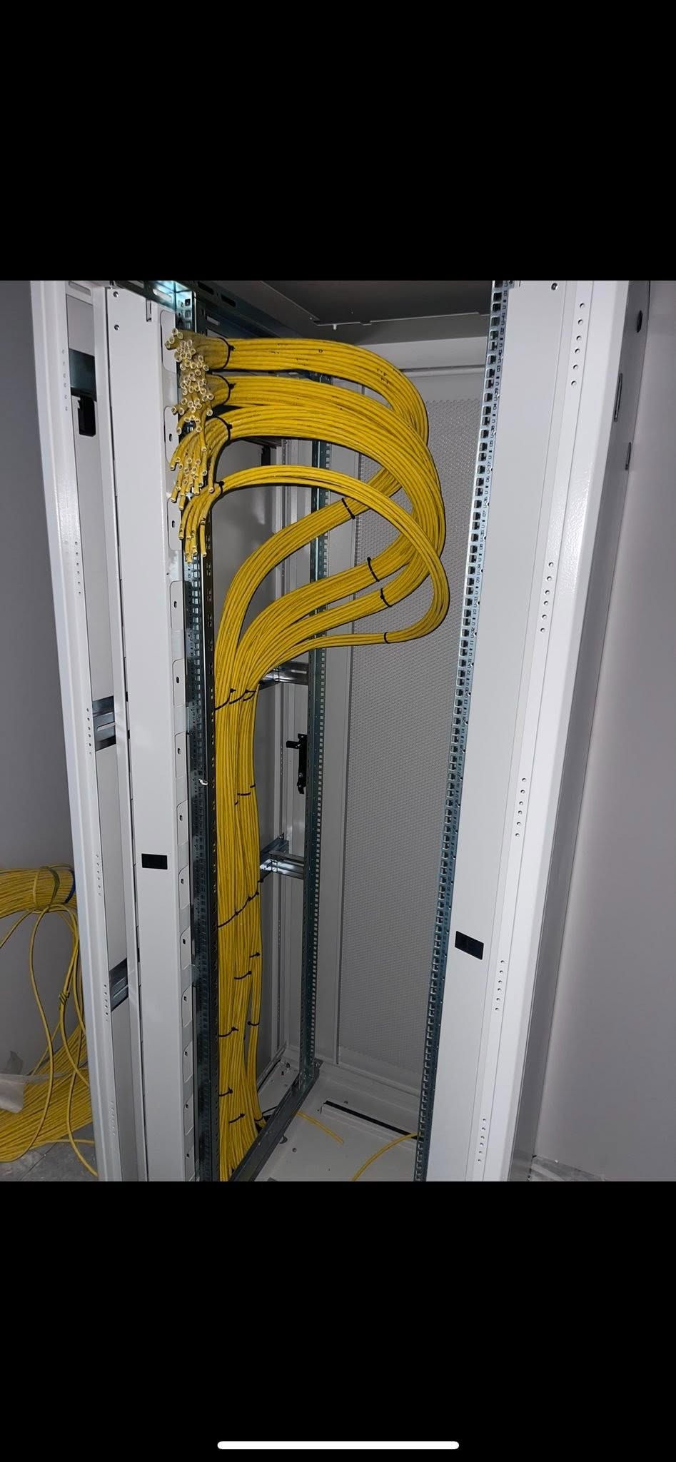 Żółte kable układane wewnątrz metalowej szafy rack, widoczne elementy mocujące i otwory wentylacyjne. Część kabli leży na podłodze obok szafy.