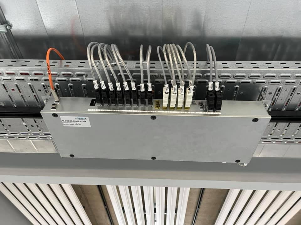 Srebrna skrzynka sterownicza SAUTER zamontowana pod sufitem z licznymi kablami podłączonymi do listwy zaciskowej na perforowanej metalowej szynie kablowej.