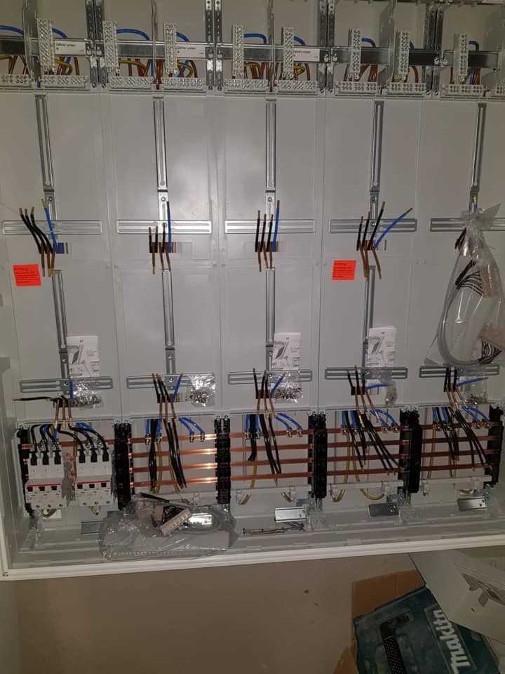 Rozdzielnia elektryczna w trakcie montażu. Widoczne szyny, kable i zabezpieczenia. Częściowo podłączone przewody. Widoczne elementy montażowe i narzędzia.