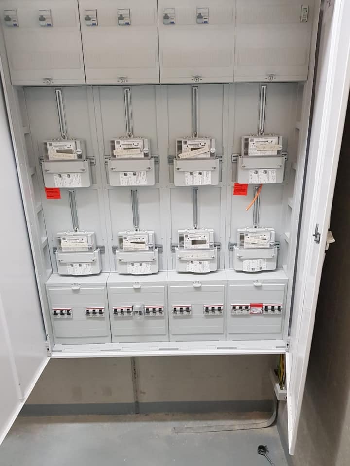 Rozdzielnica elektryczna z zamontowanymi licznikami i zabezpieczeniami, widok z otwartymi drzwiami, szara kolorystyka, równomierne oświetlenie.