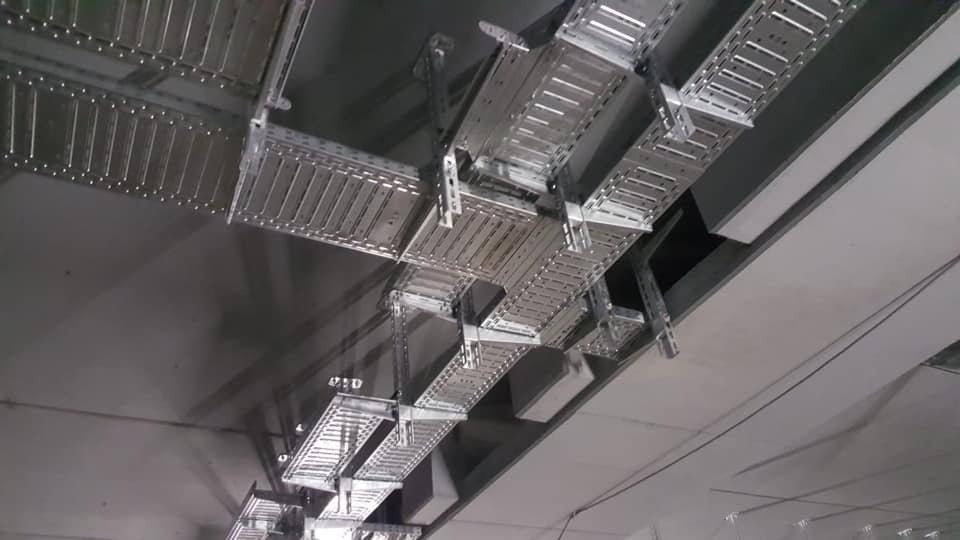 Sufit z zamontowanymi metalowymi korytkami kablowymi, widok z dołu. Konstrukcja wsparta na zawiesiach, z widocznymi elementami mocującymi i przewodami.