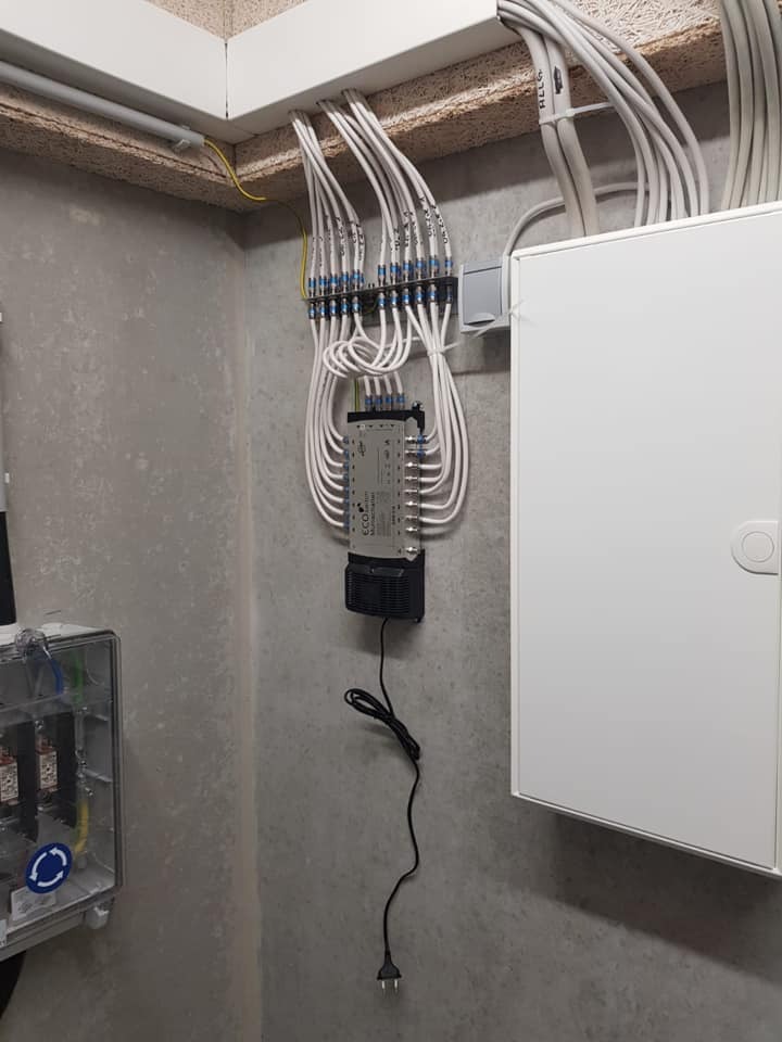 Rozdzielnica z licznymi kablami oznaczonymi numerami, zamontowana na szarej ścianie. Widoczna skrzynka elektryczna i biała szafka. Ujęcie z dołu, podkreślające złożoność instalacji.