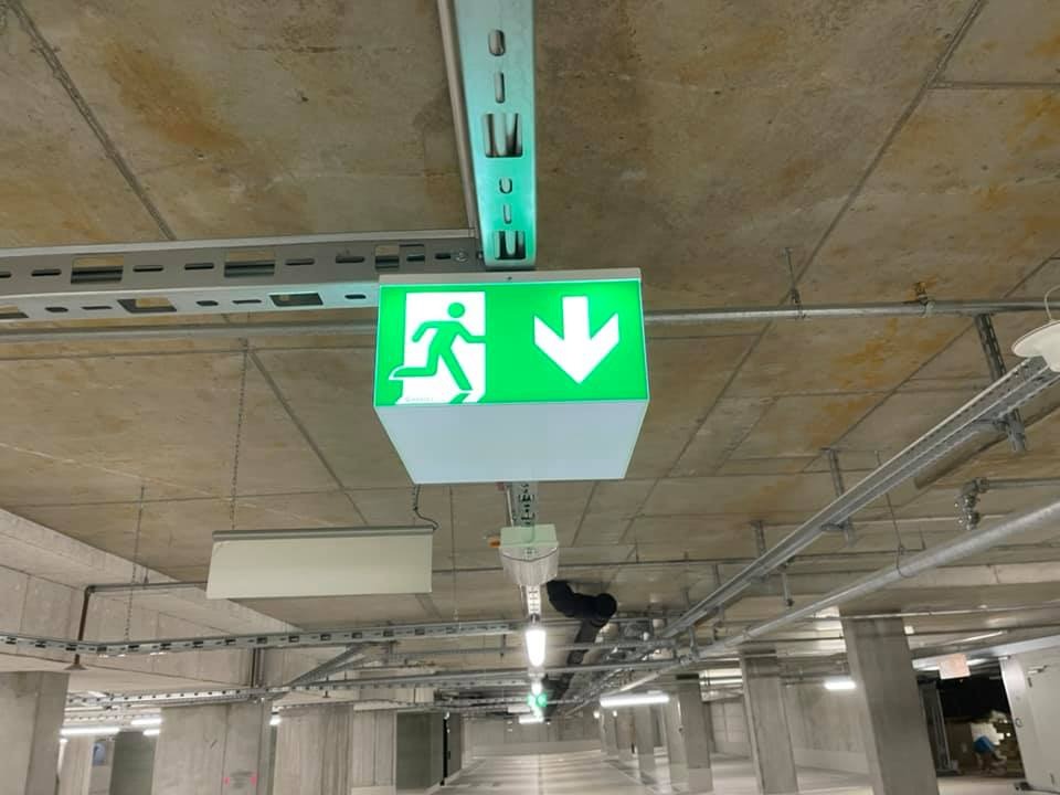 Zielony znak ewakuacyjny z piktogramem i strzałką w dół, zamontowany w garażu podziemnym w Rudce. Widoczne instalacje pod sufitem.