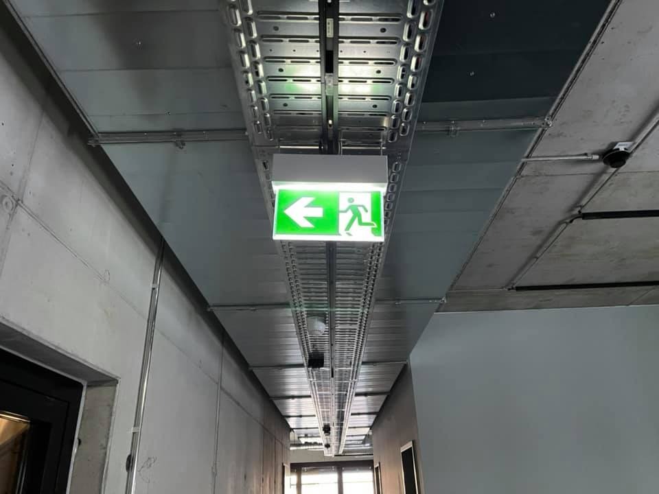 Koryto kablowe pod sufitem z betonowymi płytami i zamontowany znak ewakuacyjny ze strzałką w lewo, oświetlony na zielono.