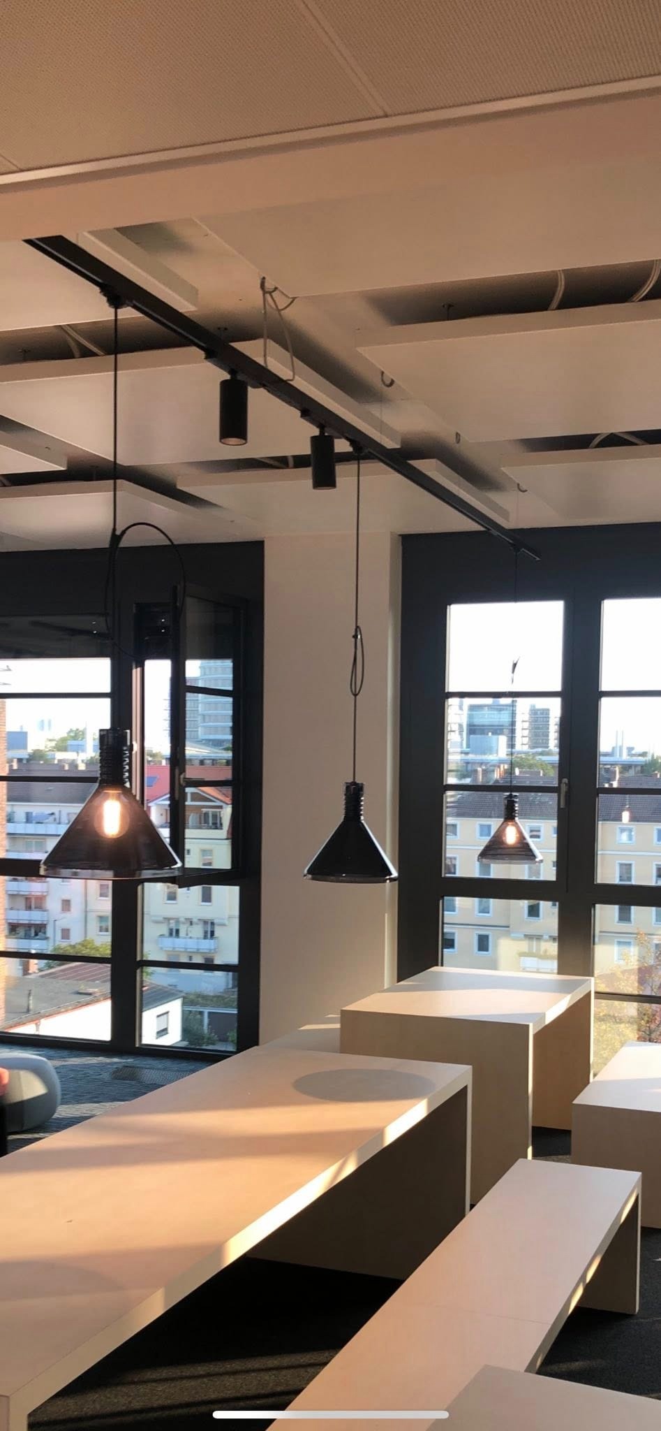 Nowoczesne lampy wiszące w biurze z widokiem na miasto. Czarne oprawy oświetlają jasne biurka. Szklane okna z czarnymi ramami w tle.