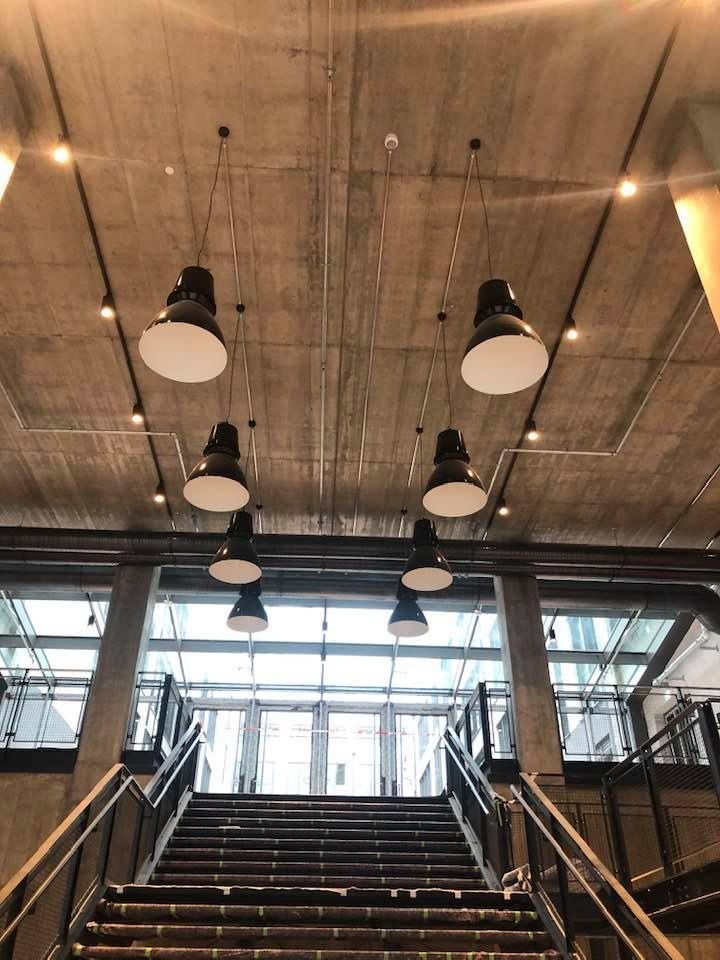 Industrialne wnętrze z betonowym sufitem, na którym zamontowano rząd czarnych lamp wiszących. W tle widoczne schody i przeszklona ściana.