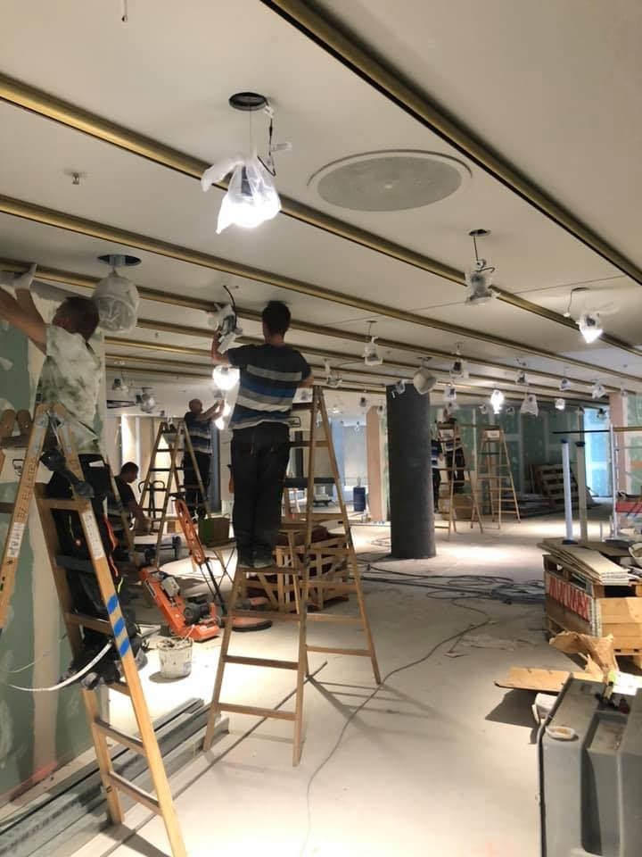 Montaż oświetlenia sufitowego w dużej przestrzeni. Elektrycy na drabinach instalują lampy w osłonach, widoczne przewody i narzędzia.