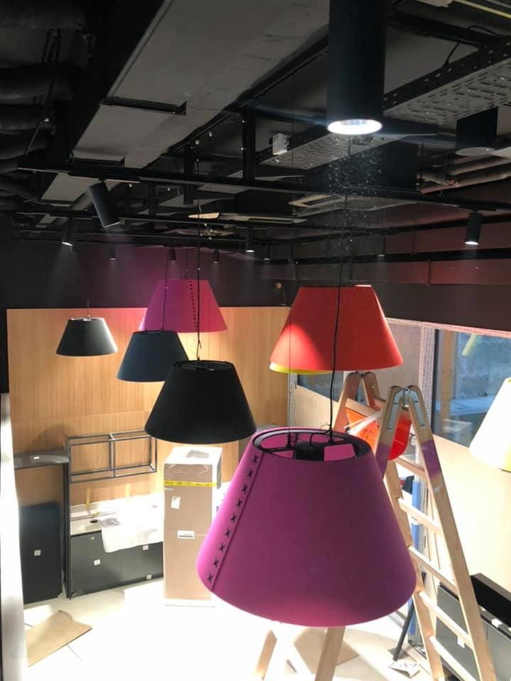 Kolorowe, stożkowe abażury wiszące na czarnych kablach, drabina oparta o ścianę, w tle elementy zabudowy meblowej i instalacje pod sufitem.