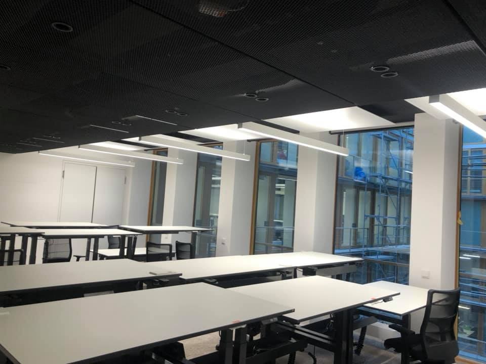 Nowoczesne biuro z rzędami biurek pod liniowymi lampami LED. Czarny sufit z perforacją. Za oknami widoczne rusztowania.