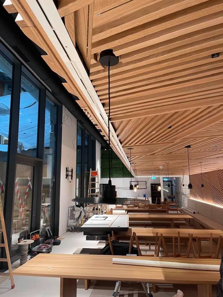 Nowoczesne wnętrze restauracji w trakcie wykańczania: drewniany sufit z oświetleniem, minimalistyczne lampy wiszące i duże okna z widokiem na zewnątrz.