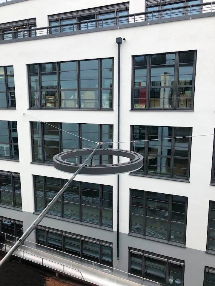 Nowoczesna, okrągła lampa wisząca zamontowana na zewnątrz budynku z dużymi oknami, widoczna z perspektywy żabiej, z przewodem biegnącym wzdłuż elewacji.