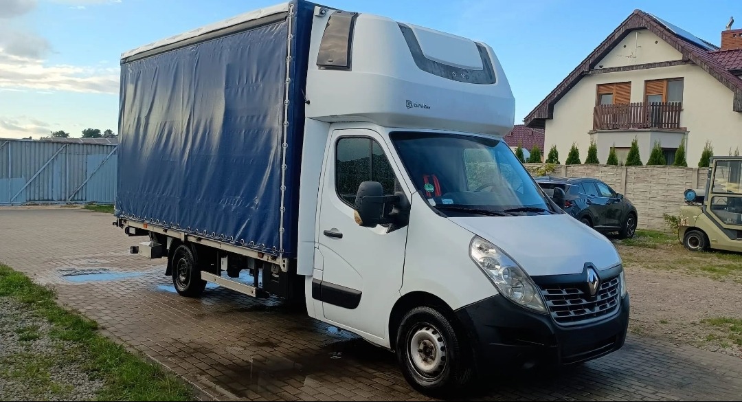 Biały Renault Master z plandeką w Morzyczynie, zaparkowany na kostce brukowej. Widoczny budynek mieszkalny w tle, błękitne niebo i zaparkowane auto osobowe.