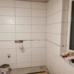 MachineBud - Białe, błyszczące płytki ceramiczne z metalowymi listwami dekoracyjnymi w trakcie montażu w kuchni lub łazience. Widoczne puszki elektryczne i wiadro z zaprawą.
