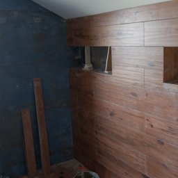 MachineBud - Łazienka w trakcie wykańczania: ciemna ściana obok okna i ściana z płytek drewnopodobnych z wnękami. Widoczne deski i resztki materiałów budowlanych.