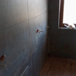 MachineBud - Ciemne płytki ścienne z wystającymi kablami i rurami w łazience, drewnopodobna podłoga, okno z brązową ramą. Faza prac wykończeniowych.