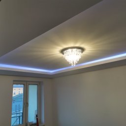 MachineBud - Wykończony sufit podwieszany z oświetleniem LED i kryształowym żyrandolem. Świeżo pomalowane ściany w jasnym odcieniu. Widok na okno i fragment budynku na zewnątrz.