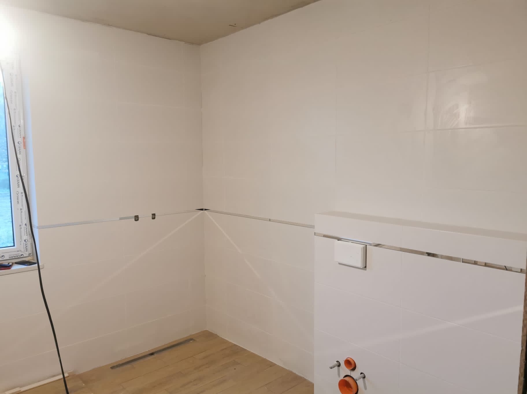 Biała łazienka w trakcie wykańczania: świeżo położone błyszczące płytki, widoczne przyłącza hydrauliczne i okno. Podłoga wyłożona panelami imitującymi drewno.