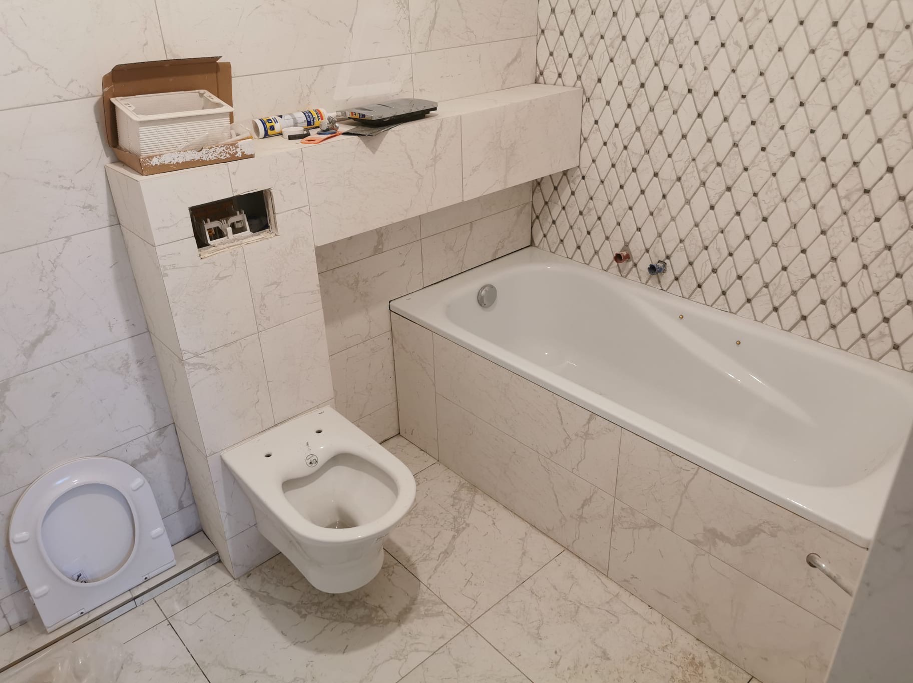 Biała łazienka w trakcie wykończenia: wanna obudowana płytkami, podwieszana toaleta bez deski. Na ścianie płytki w romby. Widoczne narzędzia i materiały budowlane.