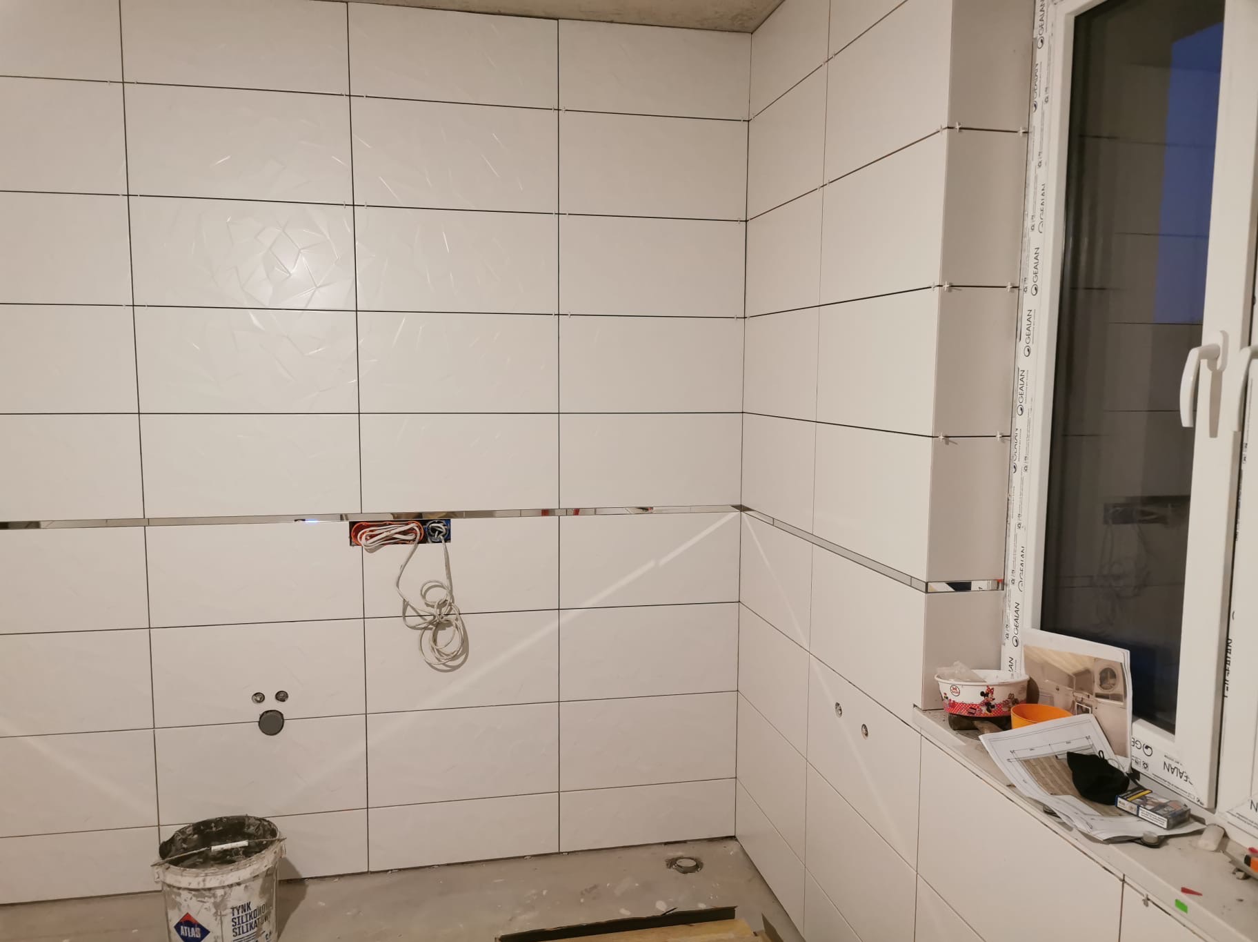 Białe, błyszczące płytki ceramiczne z metalowymi listwami dekoracyjnymi w trakcie montażu w kuchni lub łazience. Widoczne puszki elektryczne i wiadro z zaprawą.