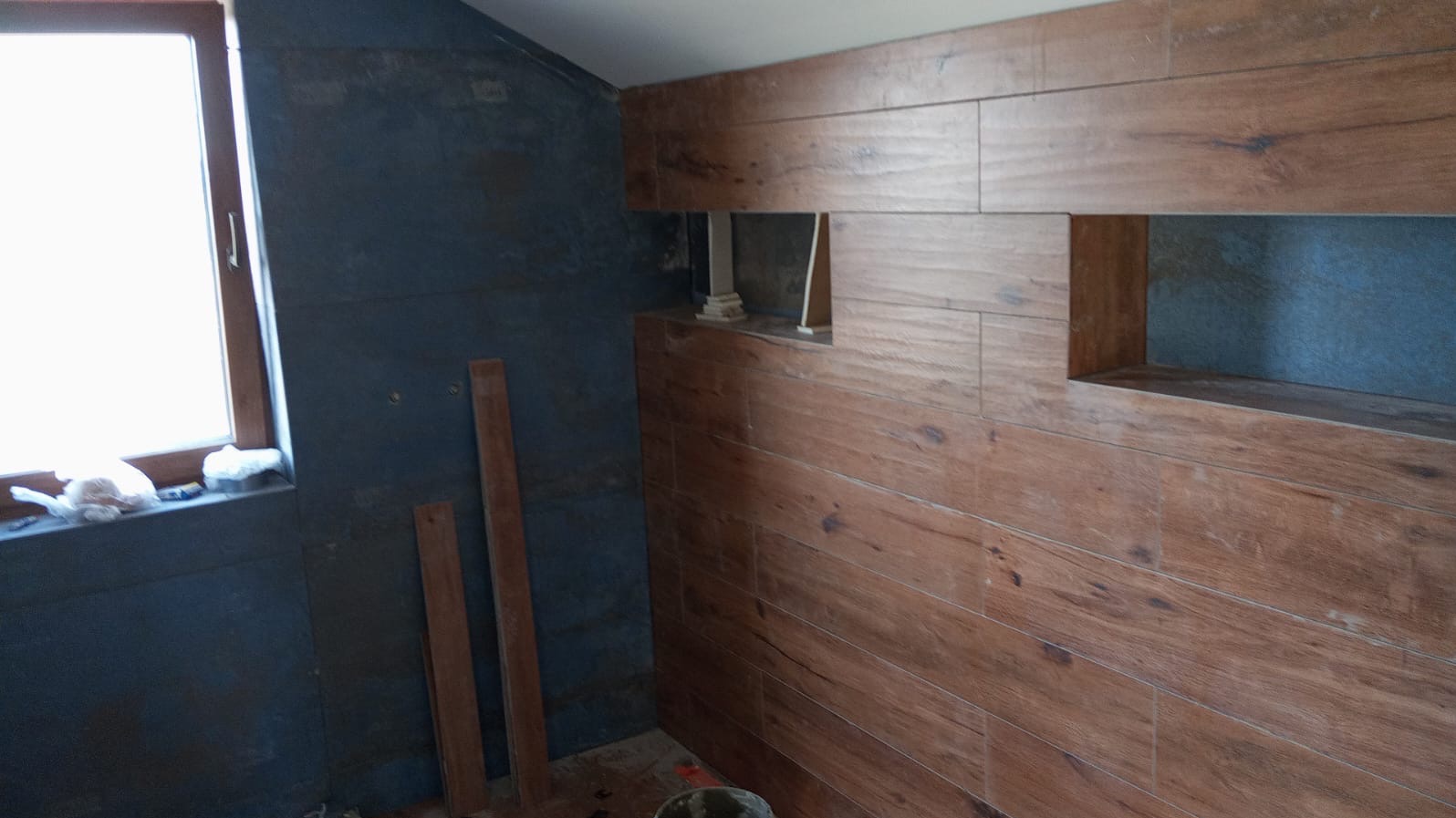 Łazienka w trakcie wykańczania: ciemna ściana obok okna i ściana z płytek drewnopodobnych z wnękami. Widoczne deski i resztki materiałów budowlanych.