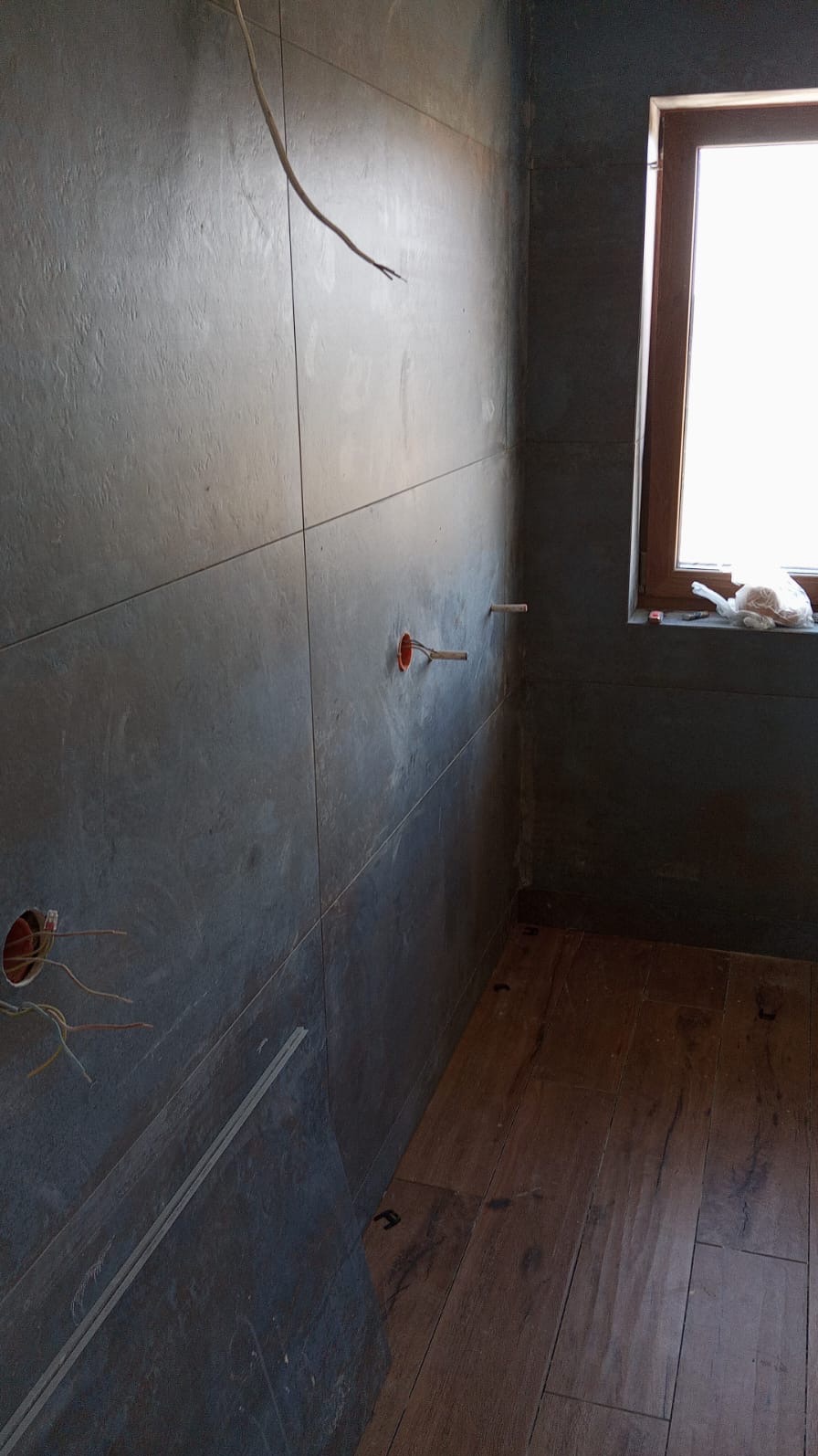 Ciemne płytki ścienne z wystającymi kablami i rurami w łazience, drewnopodobna podłoga, okno z brązową ramą. Faza prac wykończeniowych.