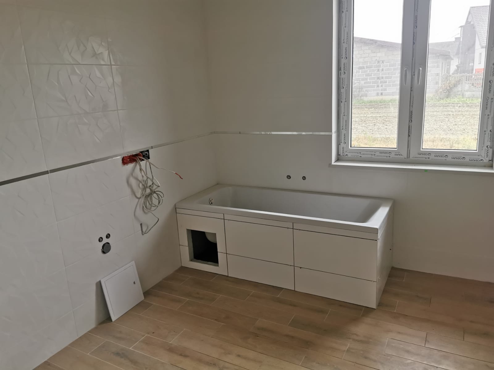 Biała, wykafelkowana łazienka z wanną w zabudowie i oknem. Widoczne niedokończone prace elektryczne i podłączenia hydrauliczne. Podłoga wyłożona panelami imitującymi drewno.
