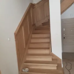 Jasne, drewniane schody z panelami ściennymi, prowadzące w górę. Widoczny fragment drzwi i sufitu. Ujęcie od dołu, podkreślające solidne wykonanie.