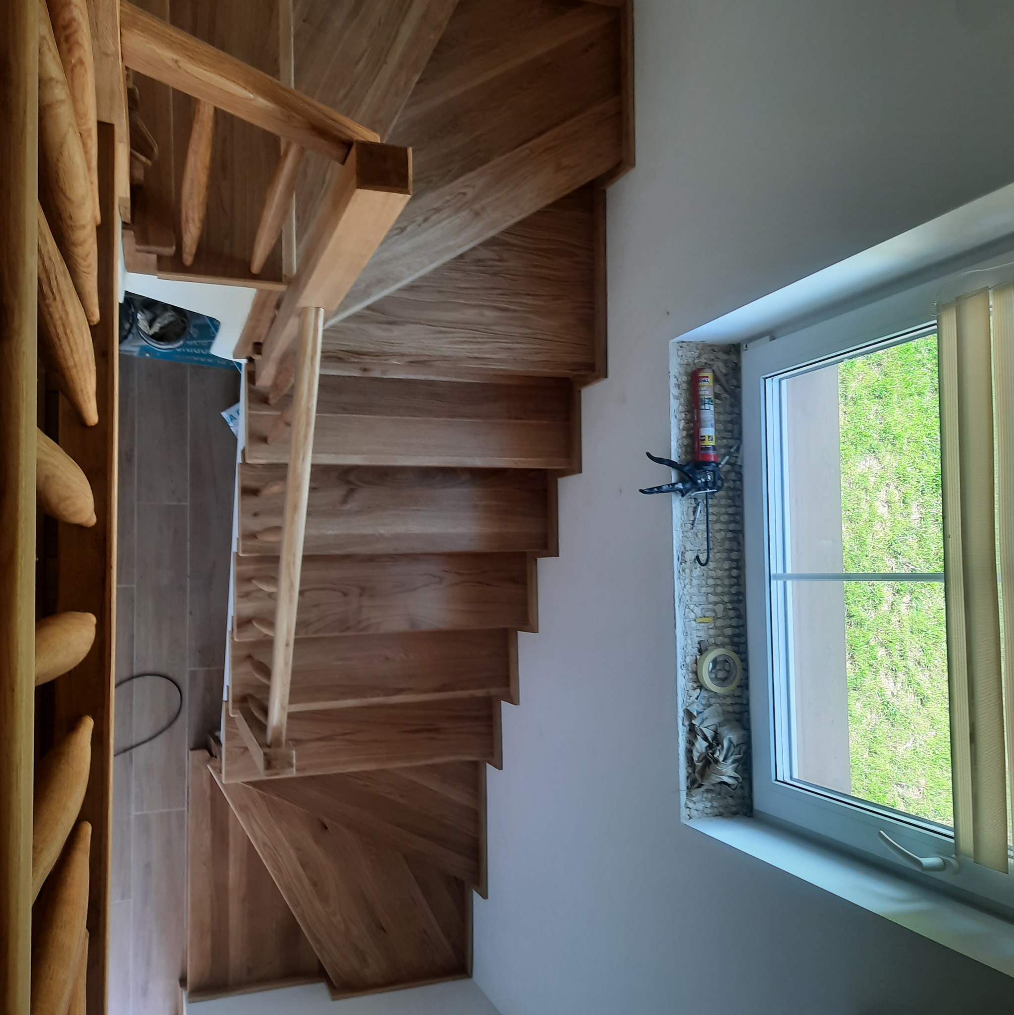 Spiralne schody z jasnego drewna, widok z góry, obok okno z widokiem na zieleń. Naturalne światło, prosta forma, minimalistyczny design.