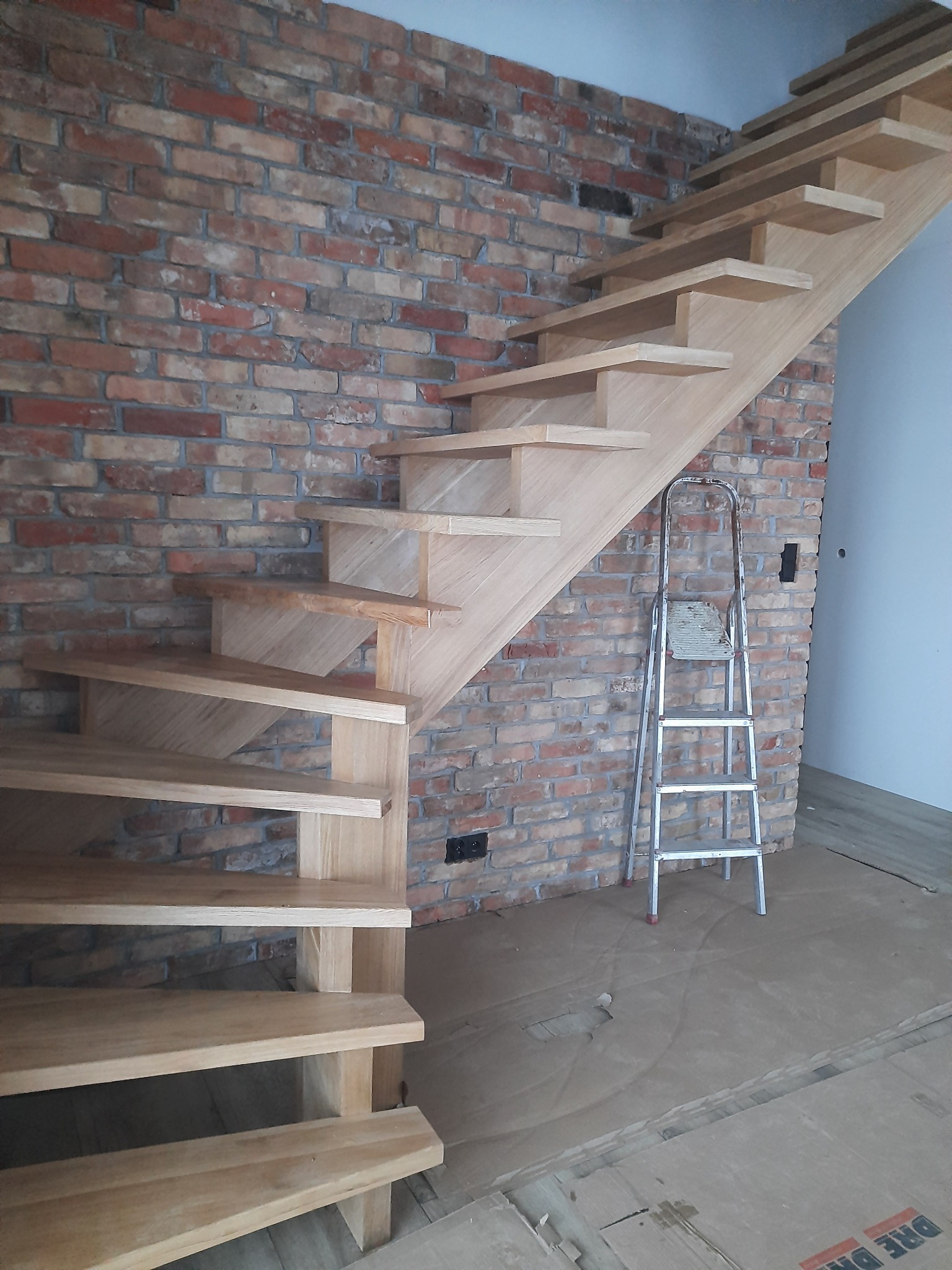 Nowoczesne, ażurowe schody z jasnego drewna, przymocowane do ściany z czerwonej cegły. Obok drabina. Podłoga zabezpieczona kartonem.