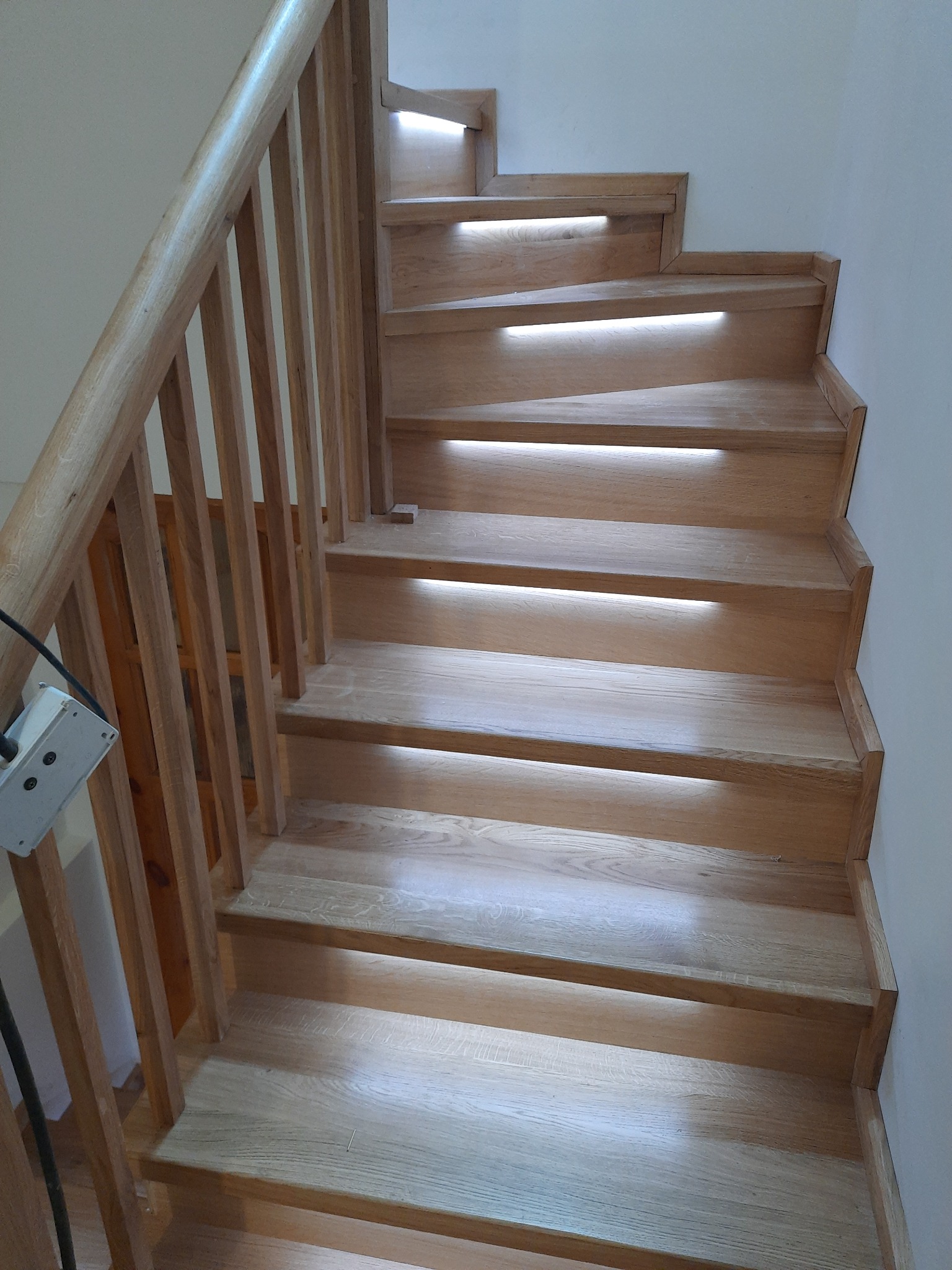 Jasne, drewniane schody z podświetleniem LED w stopniach i prostą poręczą. Widok z góry na dół, kabel zasilający widoczny po lewej stronie.